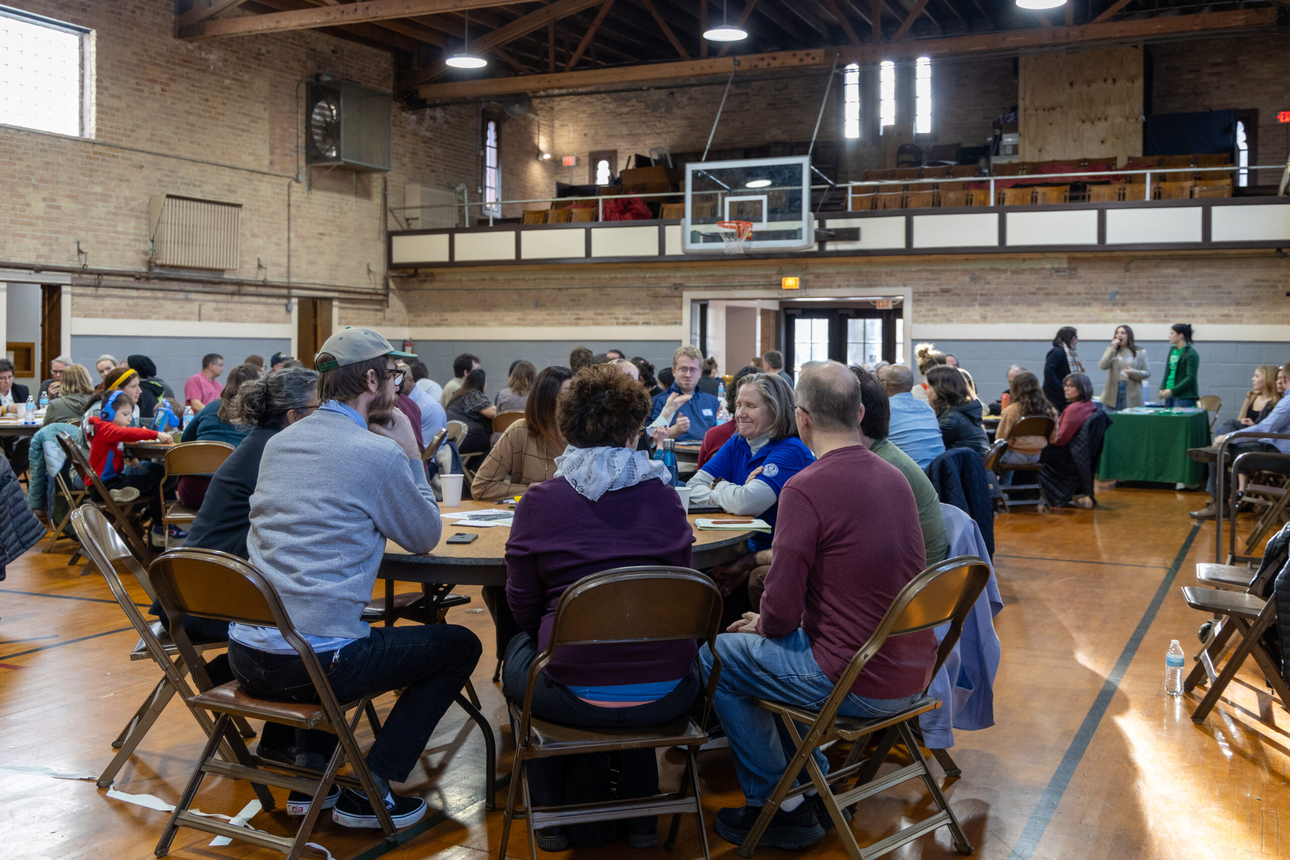 Nearly 100 people sit at tables in an indoor gym.